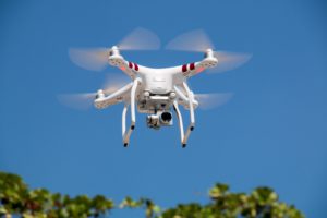 A white drone with four propellers in mid-flight against a clear blue sky. The drone is equipped with a camera and has red markings on its arms. Below, the tops of green trees can be seen, suggesting the drone is flying outdoors in a sunny environment.